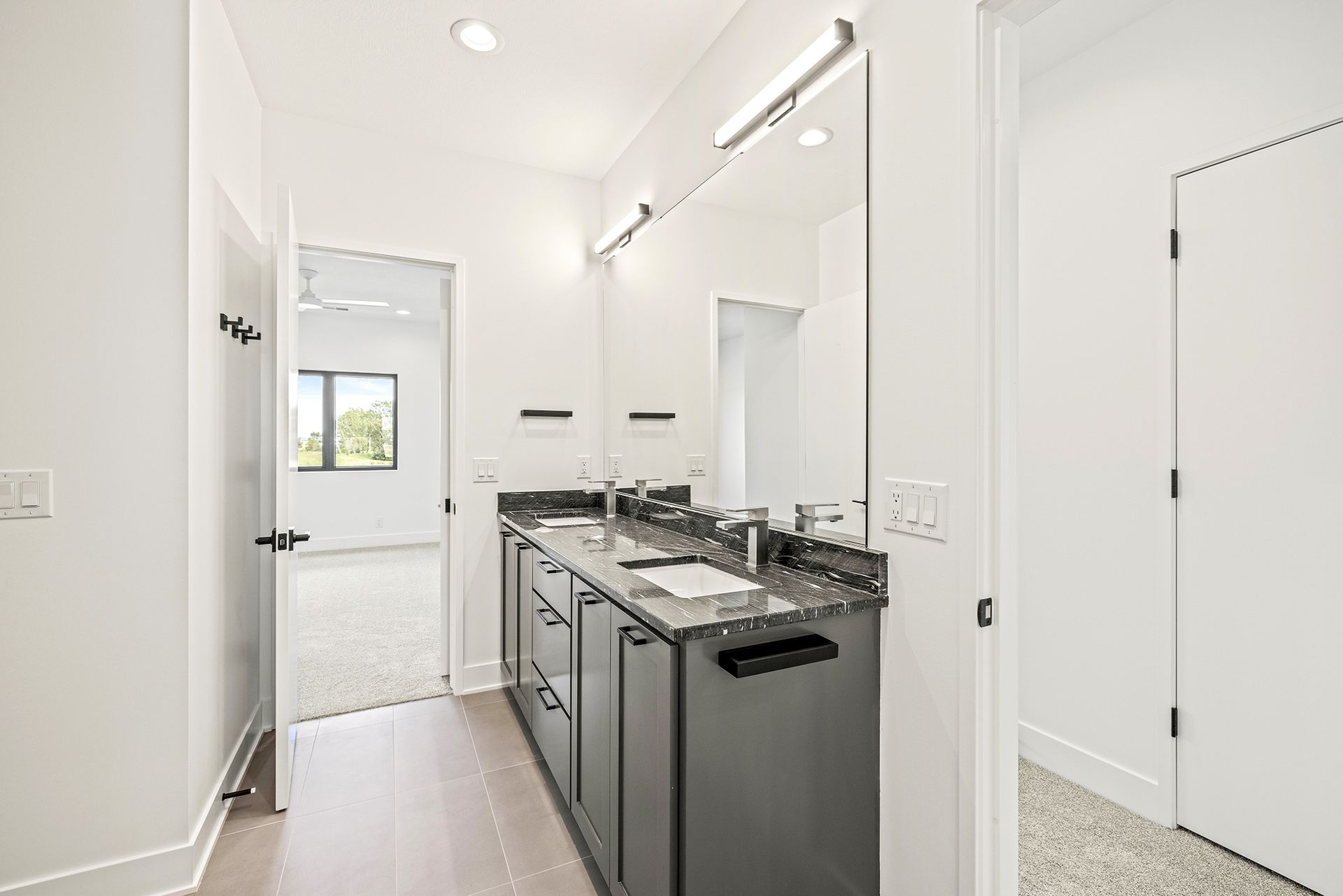 A modern bathroom featuring a dark grey vanity with a large mirror, white walls, and a door opening to a carpeted room.