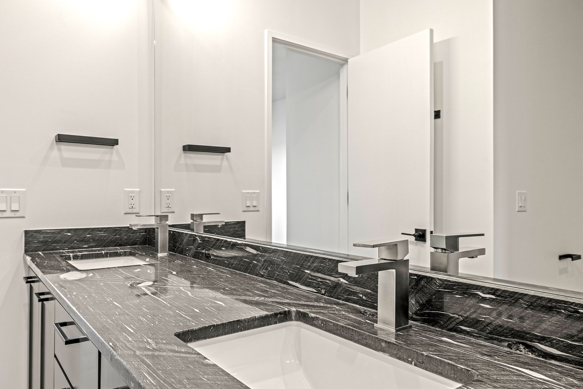 A modern bathroom vanity with a black granite countertop, two silver faucets, two white sinks, and a large mirror.