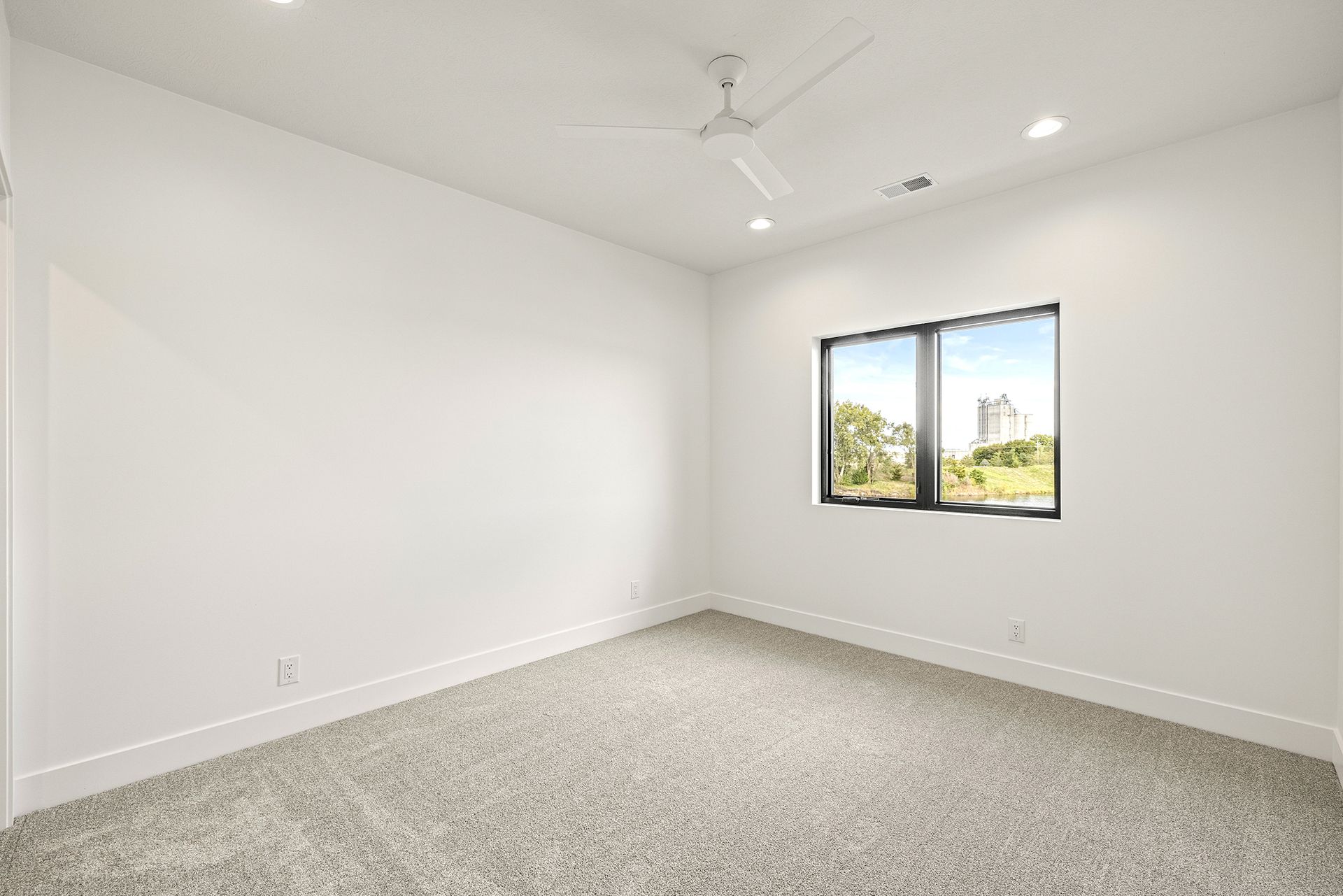 A bright, empty bedroom with white walls, gray carpet, a white ceiling fan, and a window with a view of trees.