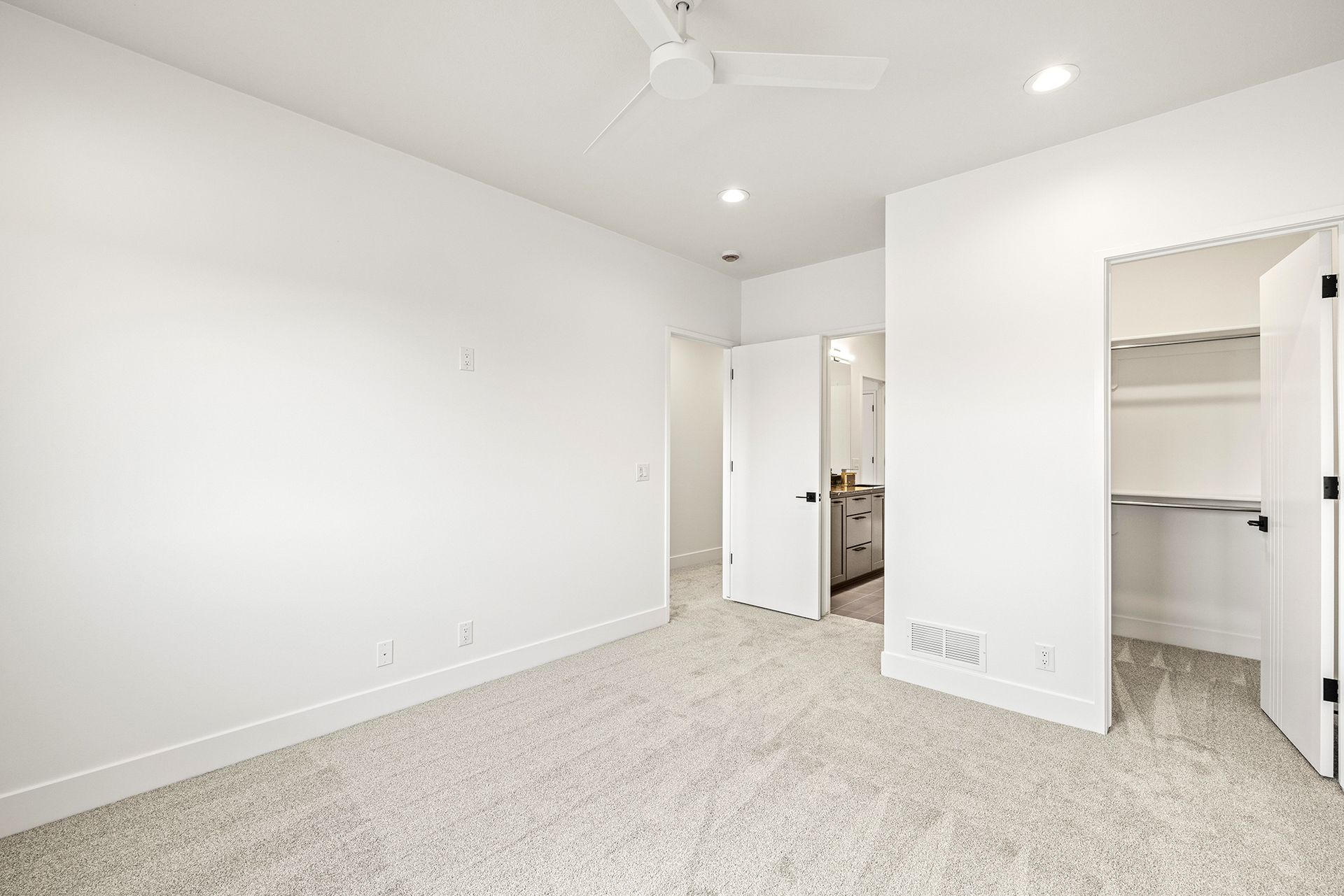 A bright, empty bedroom with white walls, light-colored carpet, a ceiling fan, and doors leading to a bathroom and closet.