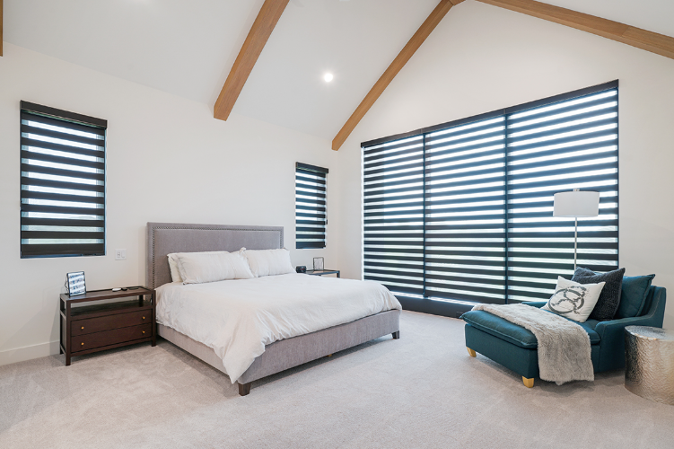 Bedroom with a bed, chaise lounge, and closed layered blinds. Beige, white, and teal colors. Wooden beams on the ceiling.