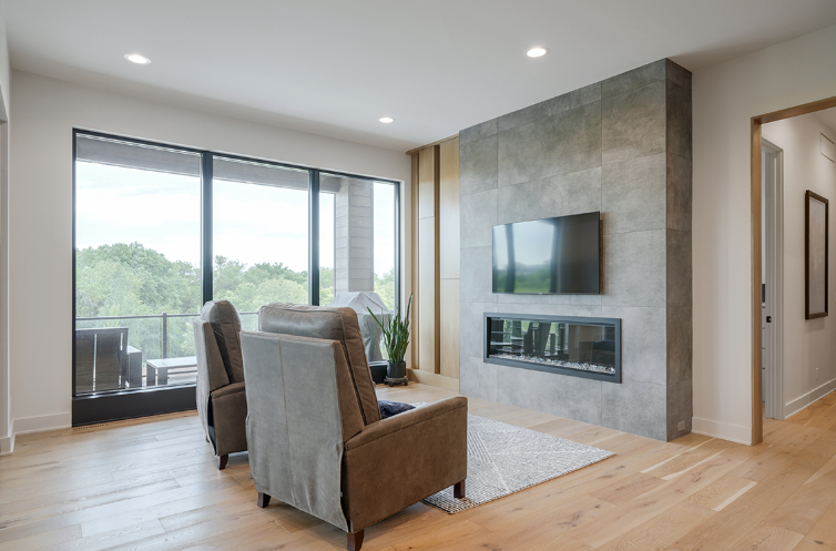 Modern living room with fireplace, TV, and two armchairs, overlooking trees through large window.
