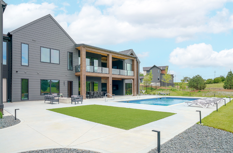 Modern house with pool and outdoor patio, blue sky. Grey siding, green grass, lounge chairs.