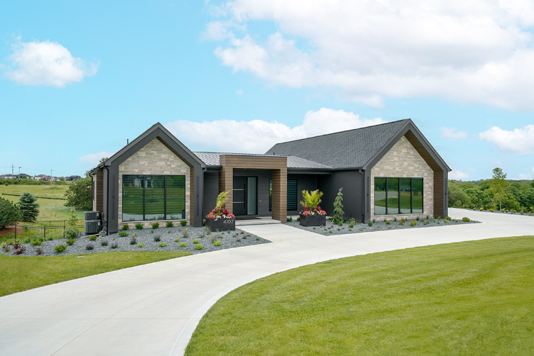 Modern, single-story house with a curved driveway. Dark siding with stone accents, large windows, and a blue sky.
