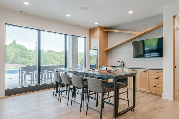Modern bar area with high-top seating, large window overlooking a pool, built-in cabinets, and a mounted TV.