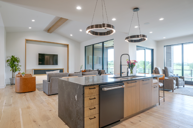 Modern kitchen with island, living room with gray couch, and large windows.