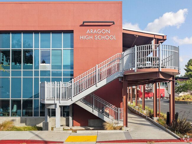 Aragon High School building exterior with red walls, large windows, and a metal staircase under a sunny sky.