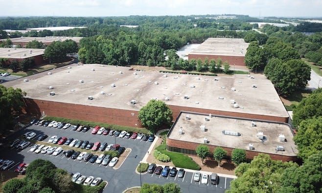 Large brick buildings with flat roofs, surrounded by trees and a parking lot full of cars.