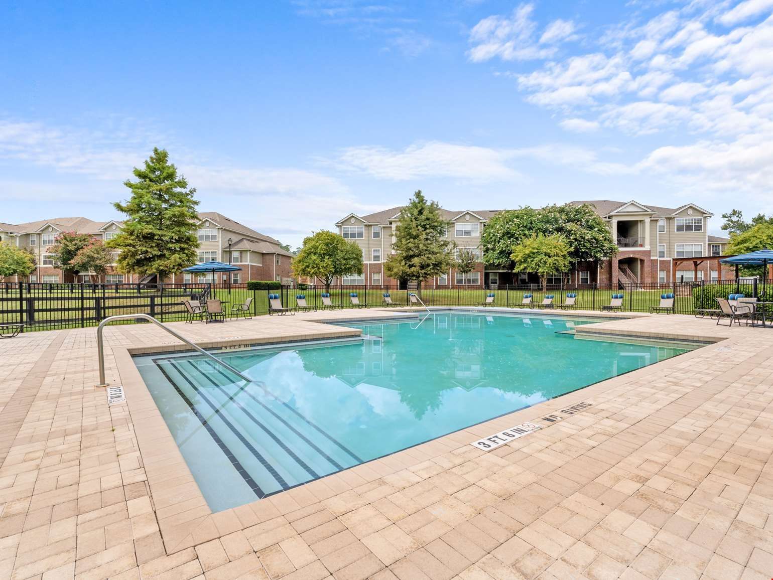 Pool surrounded by brick patio, with apartment buildings and trees under a blue sky.