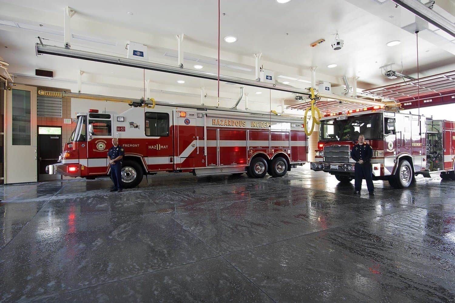 Fire station with two red and white fire trucks. A firefighter stands by one. Water on the floor.