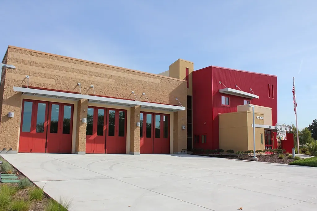 Fire station with red doors, tan brick facade, and a red tower on a sunny day.