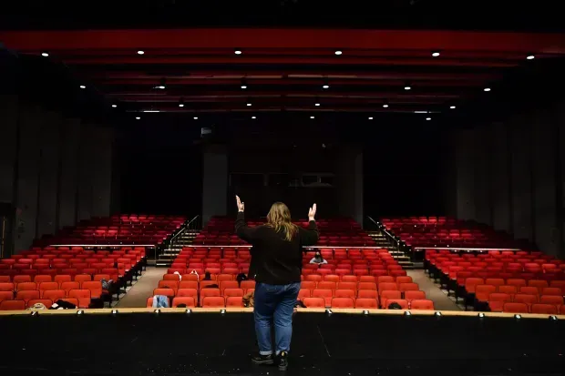 Person on stage with arms raised faces red seats in an empty theater.