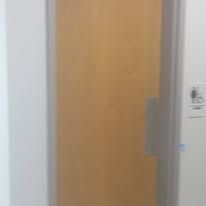 A light wood door with a long silver handle, next to a white wall with a handicap symbol.