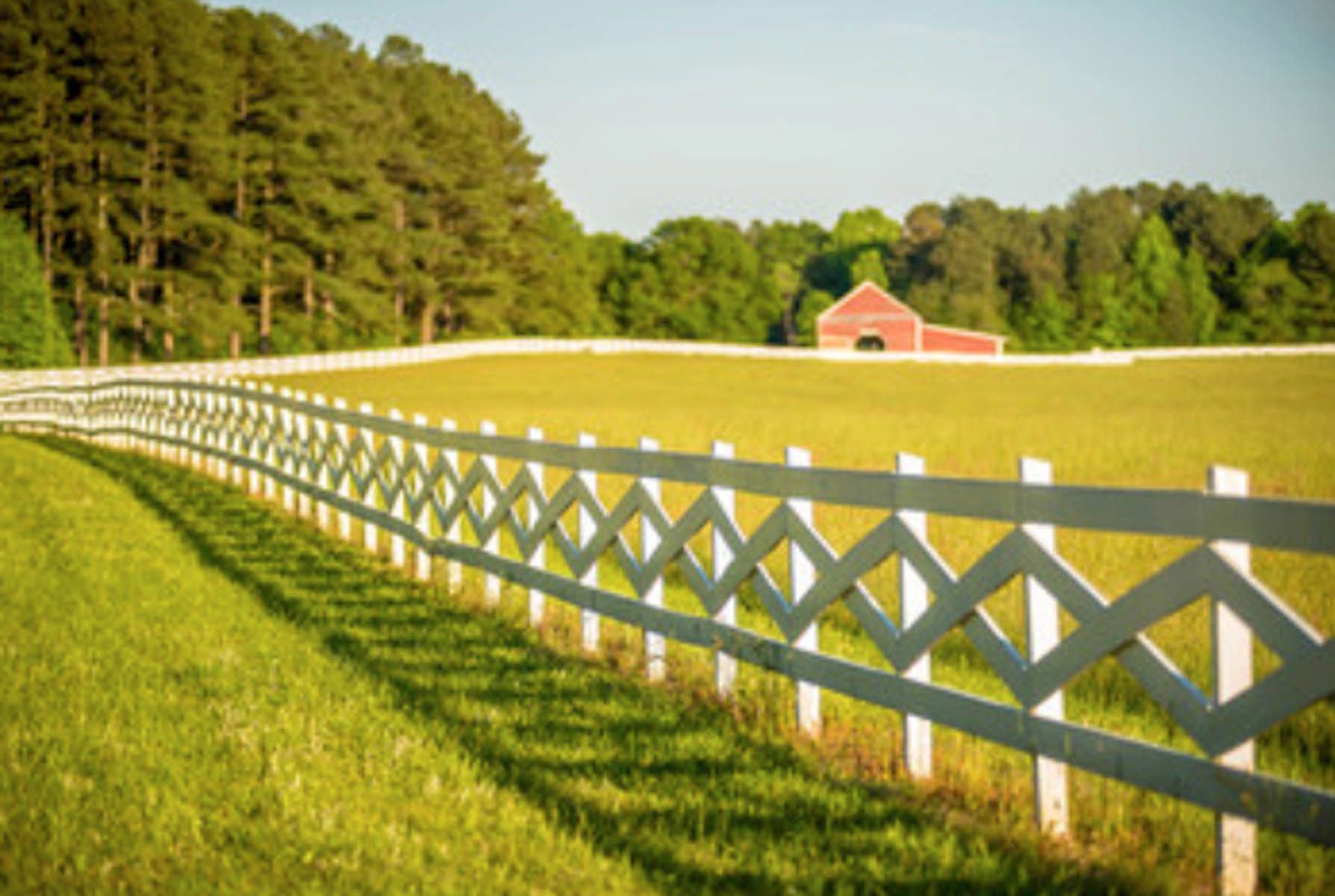 A chain-link fence in the foreground overlooks a grassy field with a military vehicle and trees under a bright sky.