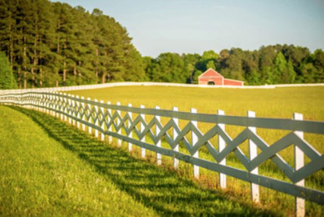 A chain-link fence in the foreground overlooks a grassy field with a military vehicle and trees under a bright sky.