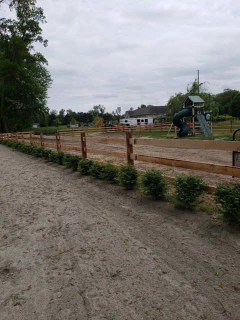 A wooden split-rail fence separates a dirt pathway from a grassy yard featuring a playground set and distant buildings.