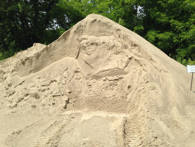 Large pile of tan sand with tire tracks; green trees in the background.