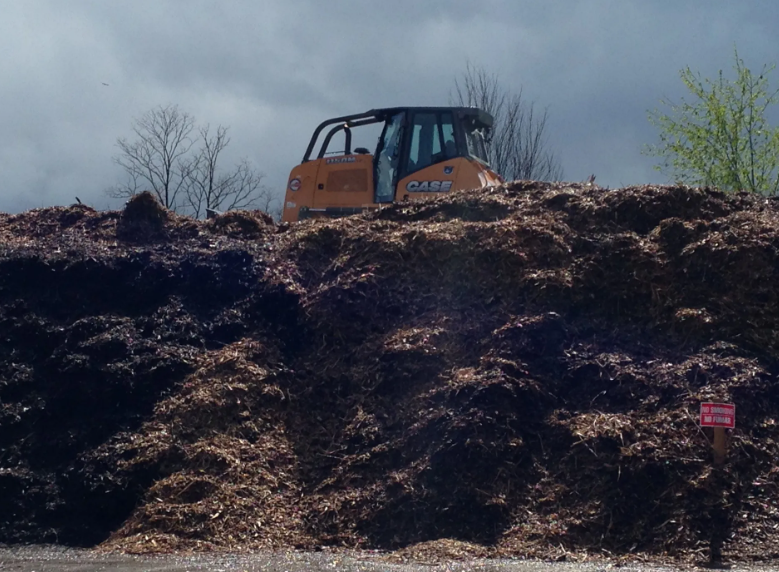 Orange skid steer on a large pile of wood chips, under a cloudy sky.