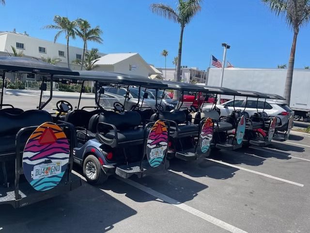 Row of colorful parked golf carts in a sunny outdoor lot with palm trees and buildings in the background