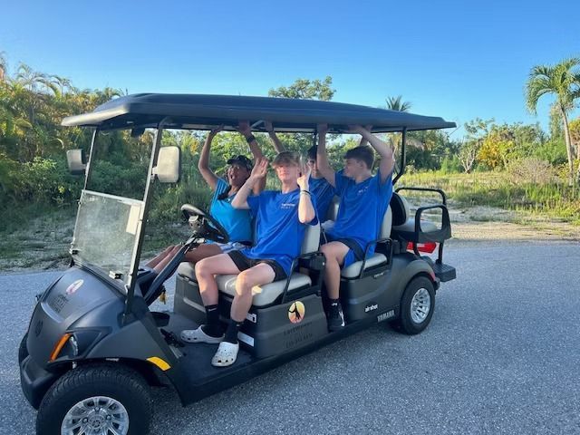 Four people in blue shirts ride in a golf cart on a sunny road, smiling and waving.