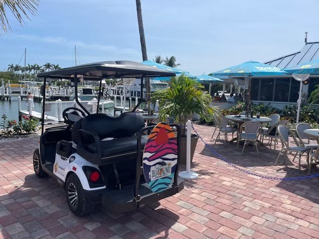 Golf cart parked on a sunny marina patio beside outdoor tables and blue umbrellas