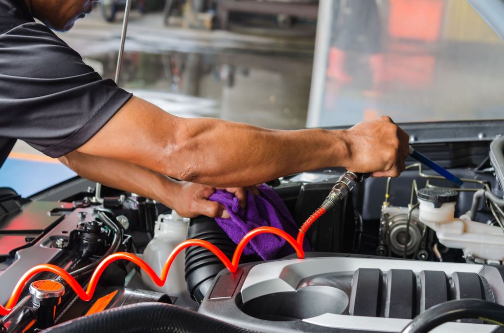 Mechanic cleaning an engine bay with an air hose and purple cloth, with red coiled tubing visible