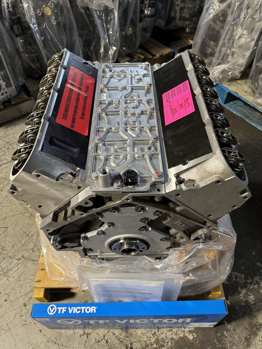Engine block on a pallet, silver with red and pink labels, in an industrial warehouse setting