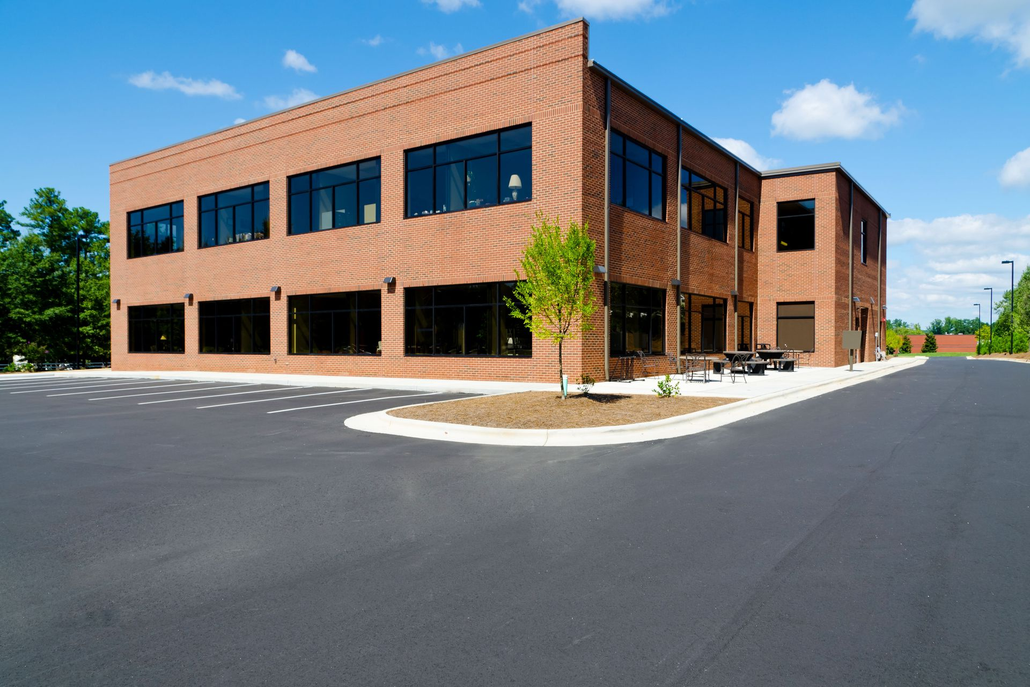 Brick office building with dark windows and a large empty parking lot on a sunny day.