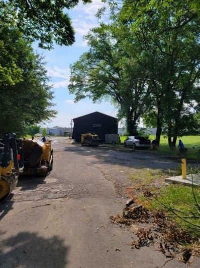 Gravel driveway with tractors leading to a dark barn under tall green trees on a sunny day.