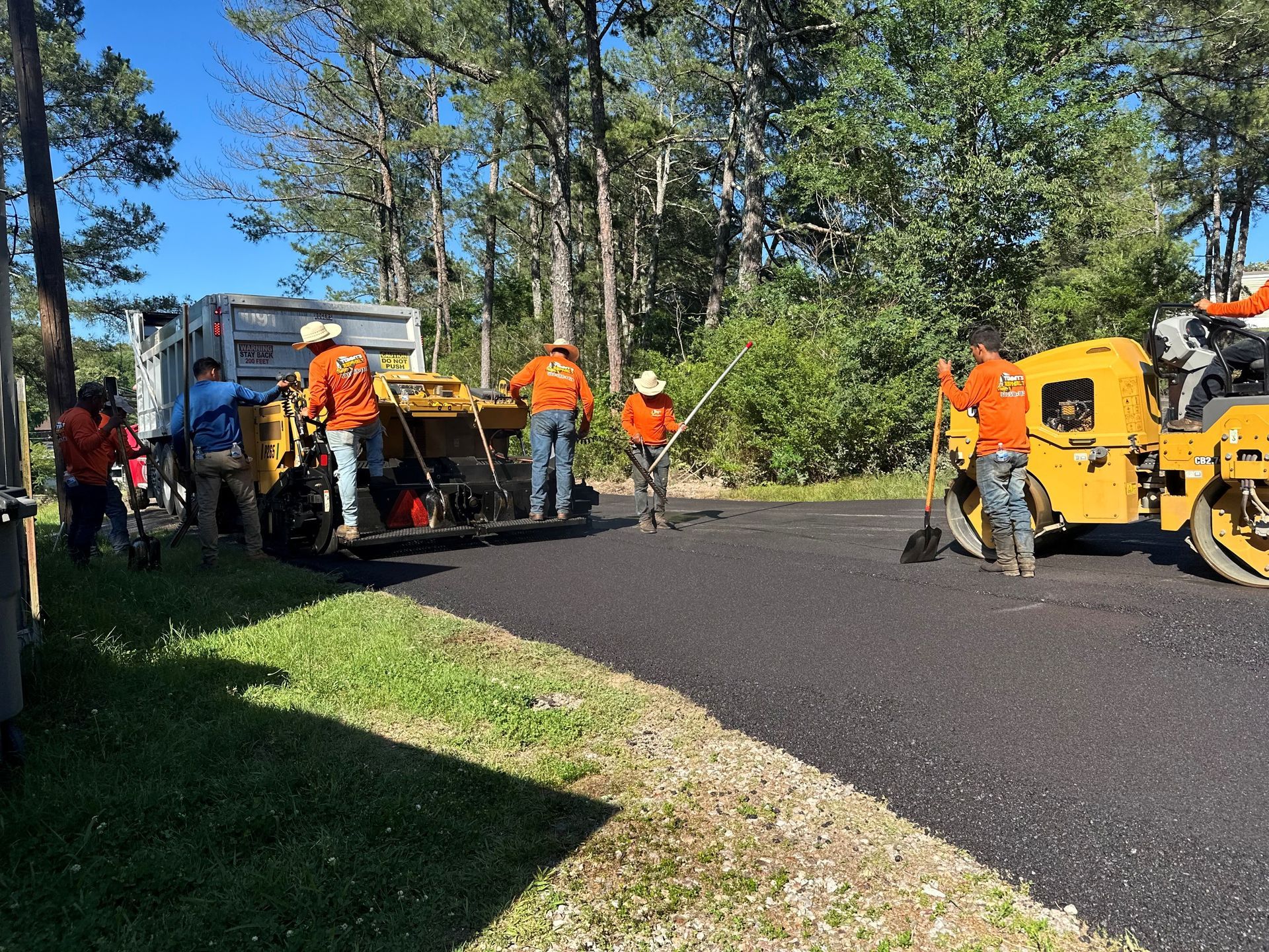 Workers in orange safety gear paving a road beside heavy machinery in a wooded area.