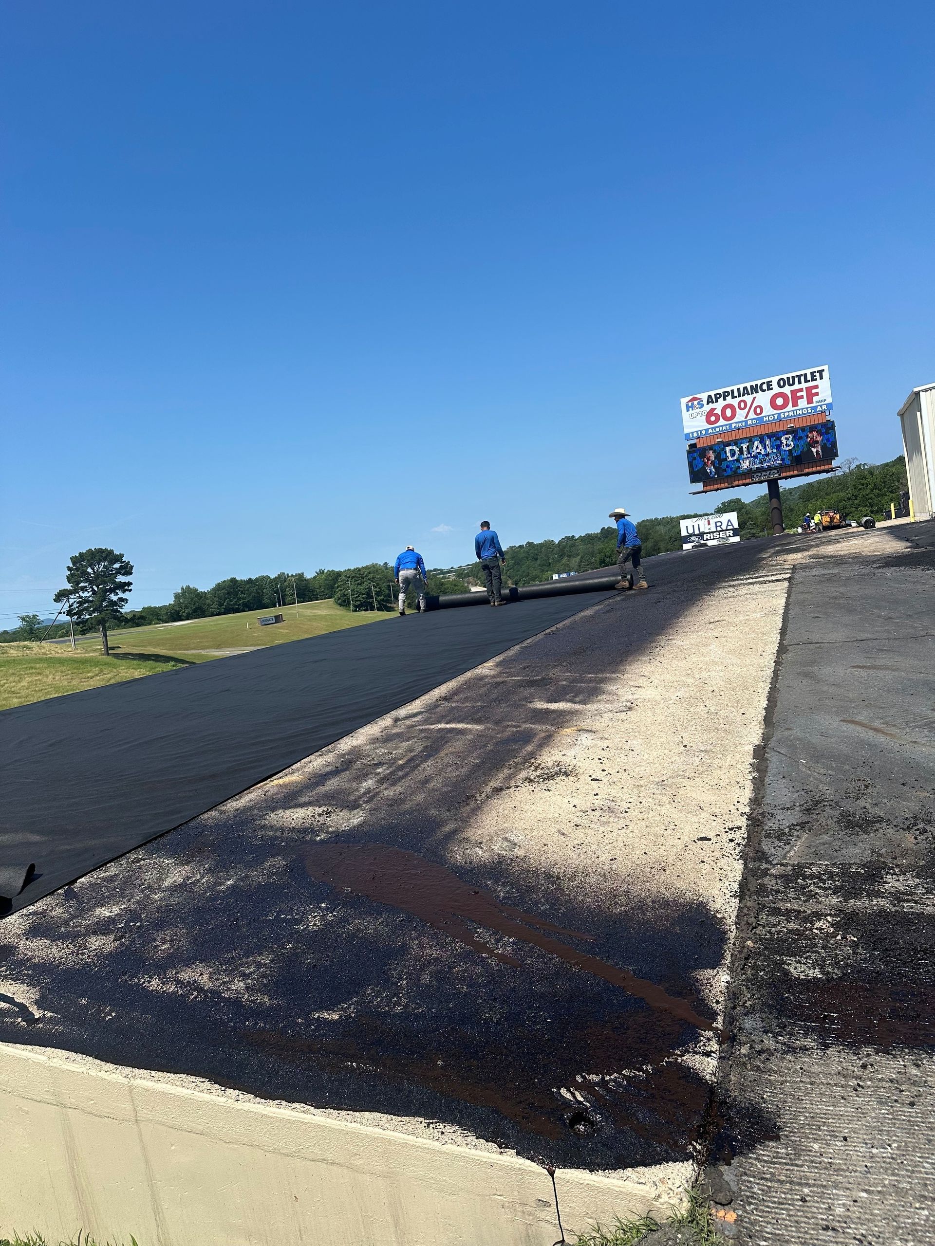 Freshly paved road with workers under a blue sky and a roadside sign in the distance.