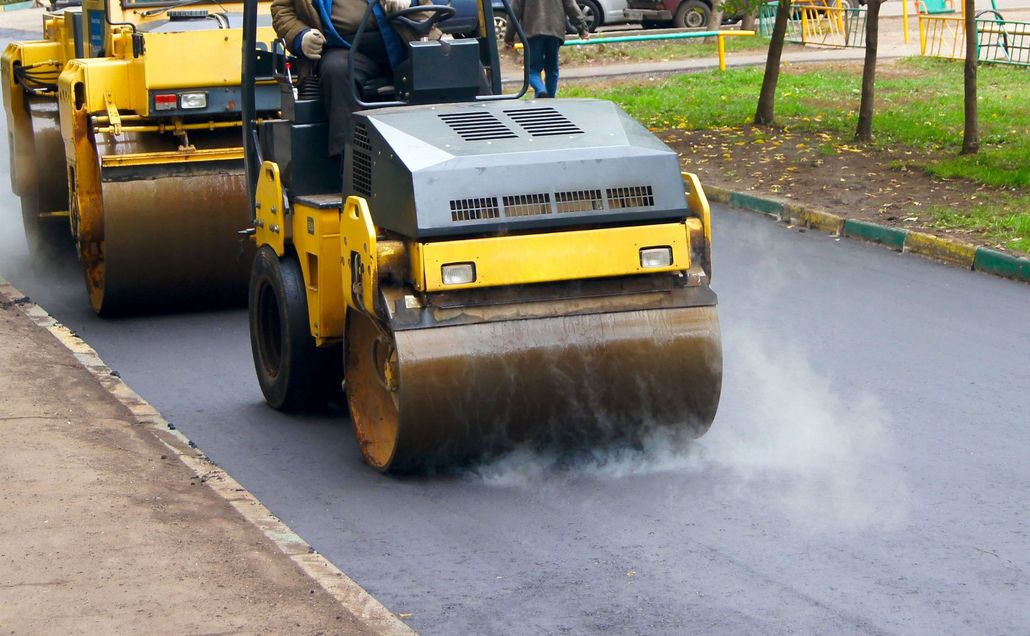 Yellow road roller compacting fresh asphalt on a street, with steam rising behind it.