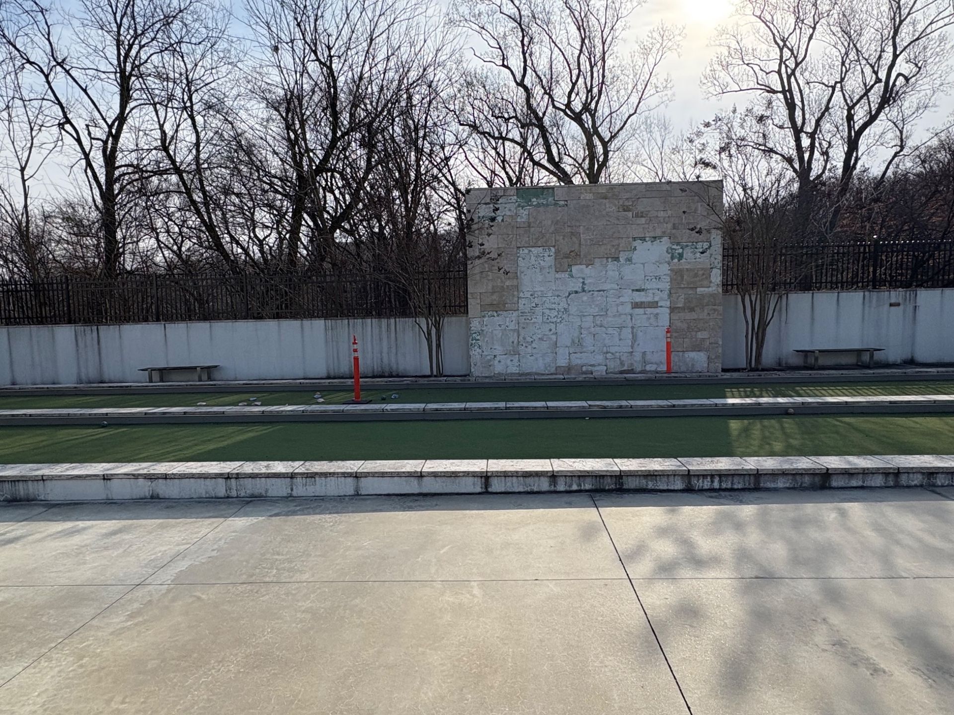 Concrete surface in front of a green field, a brick wall, and bare trees under a bright sky.