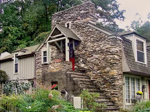 Stone house with steep stairs leading to a covered entrance. Lush garden and trees surround the structure.