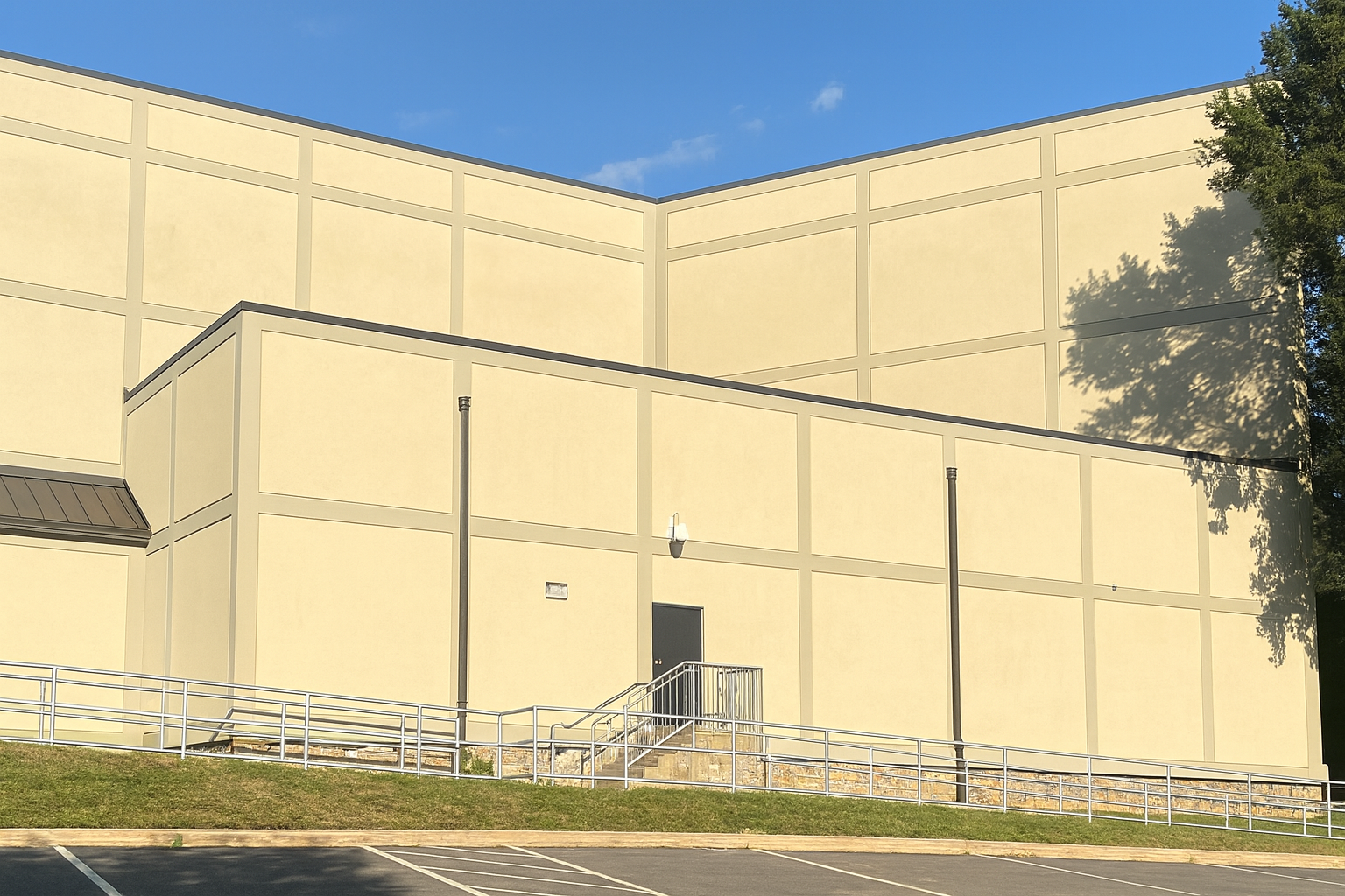 Beige building with a dark roof and door. A metal railing is along a small set of stairs. Green grass and blue sky.