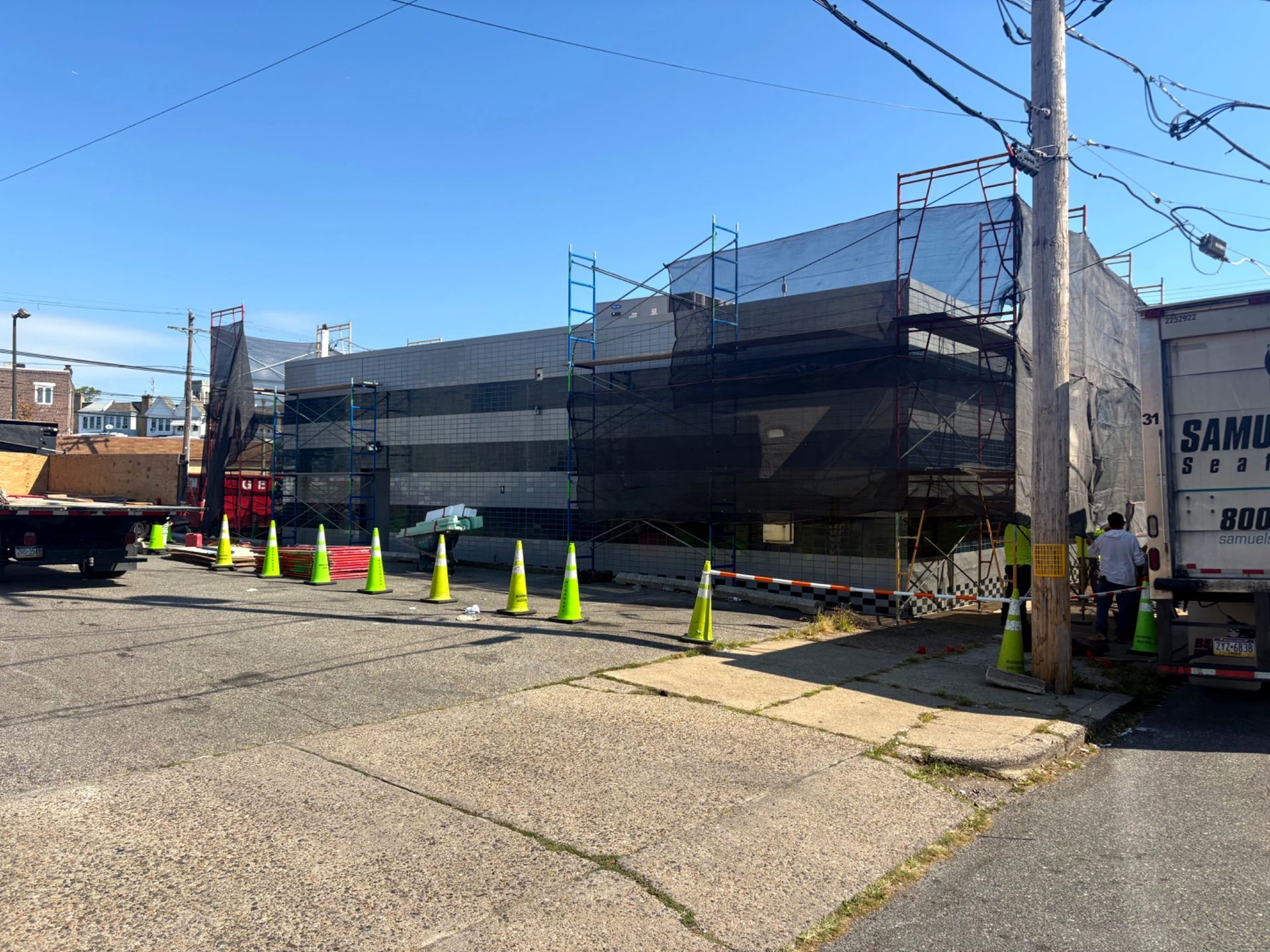 Building under construction, covered in scaffolding and black netting. Cones line the street. A truck is parked to the right.