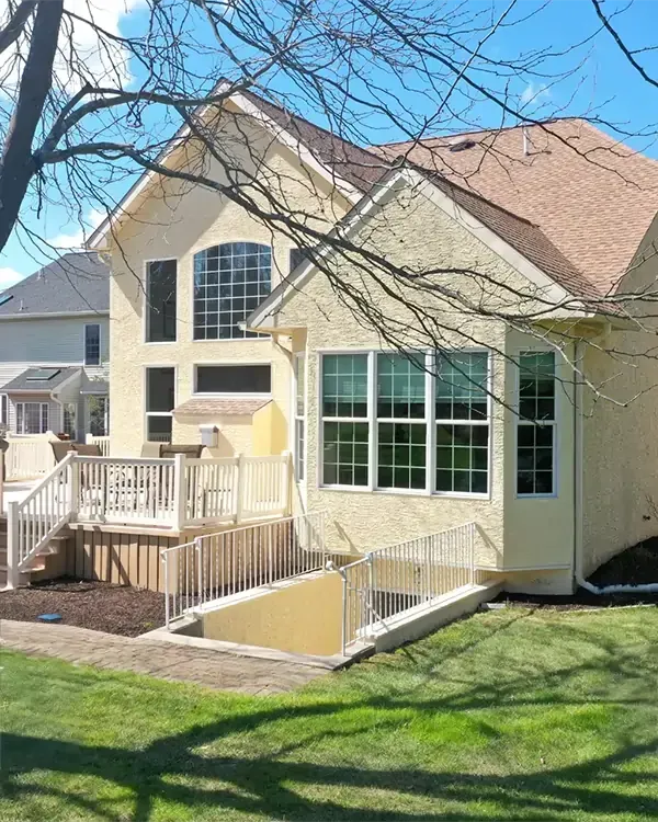 Beige house with a deck and basement entrance, surrounded by a grassy yard.