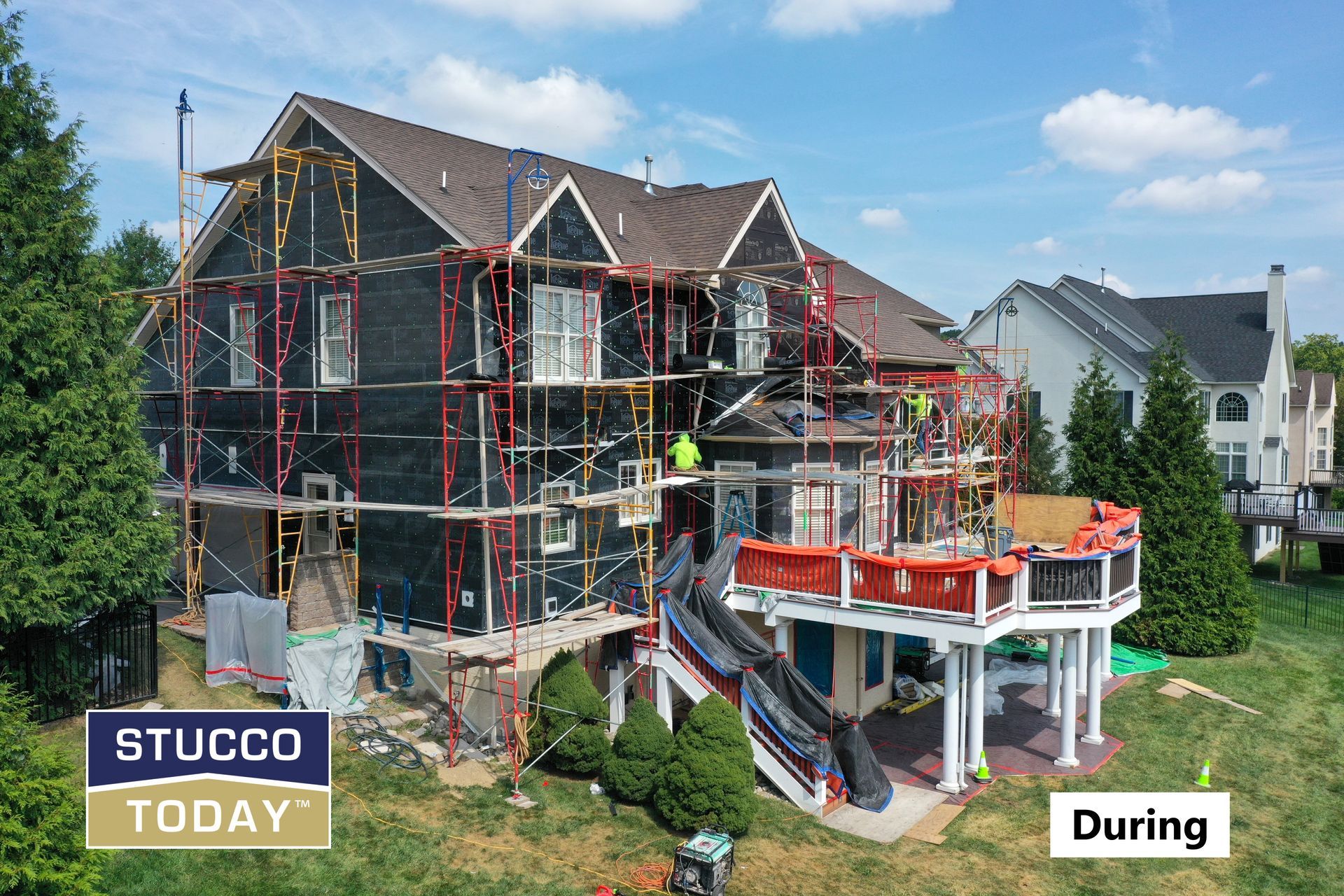 House under construction, scaffolding, black siding, workers, brown roof, white deck, green lawn, clear sky.