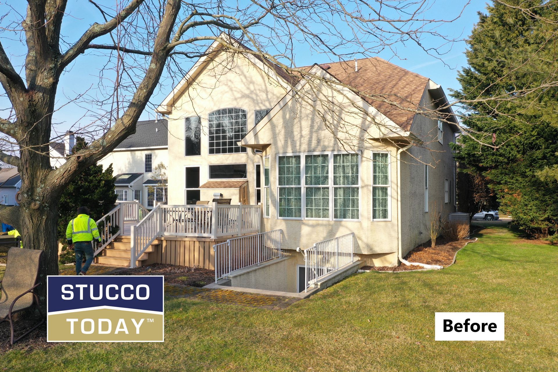 House with stucco siding before renovation, surrounded by lawn and trees. A worker in a vest is walking.