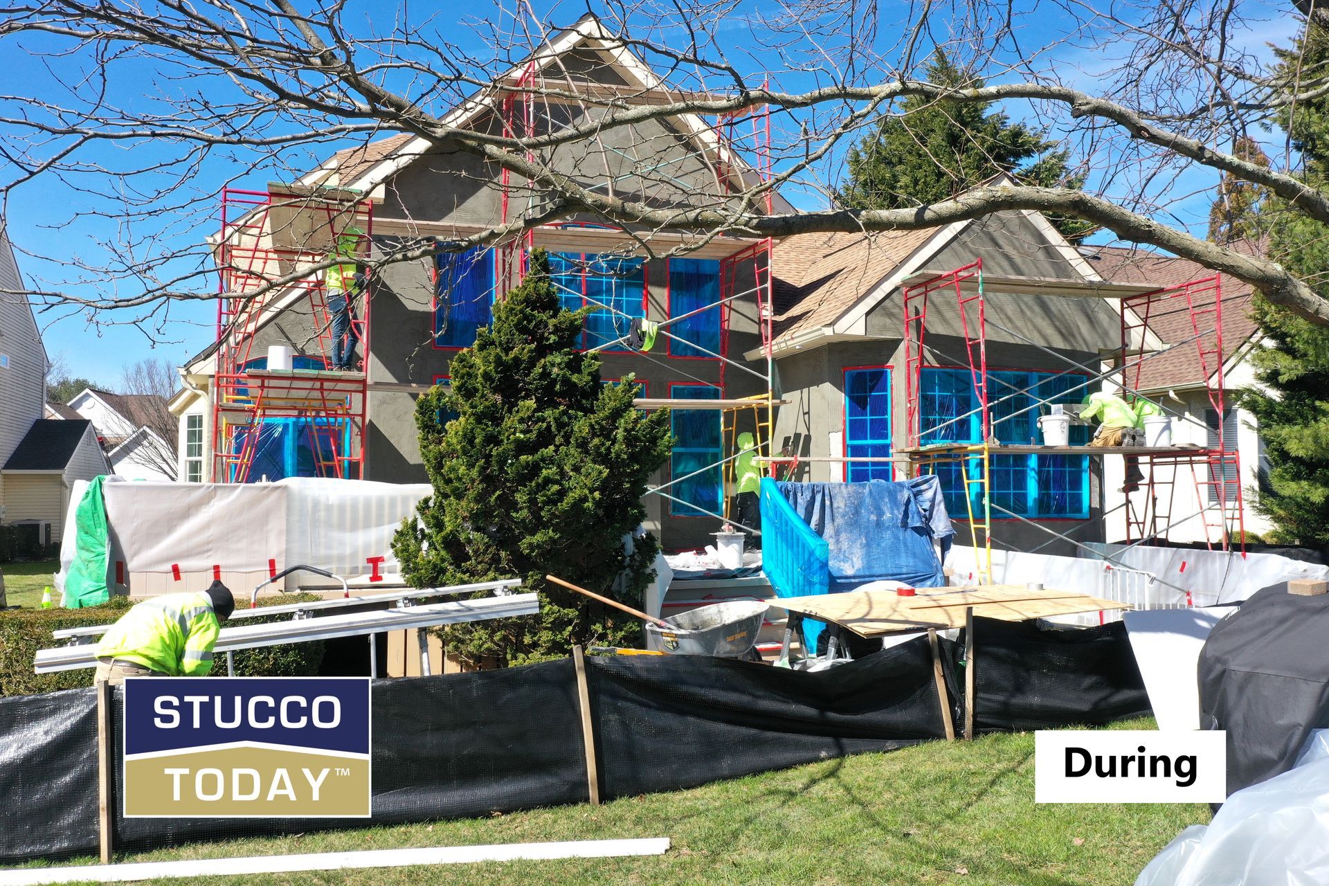 House under construction with scaffolding; workers applying stucco. Blue windows, green grass, and trees.