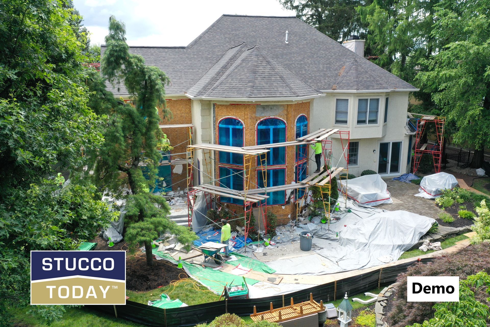 House under renovation, scaffolding, workers, stucco application. Exterior view, surrounded by trees and debris.