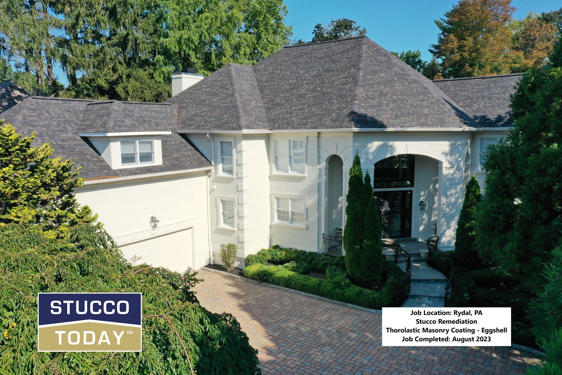 Exterior view of a large, two-story stucco house with a dark gray roof. Lush greenery and a driveway are visible.