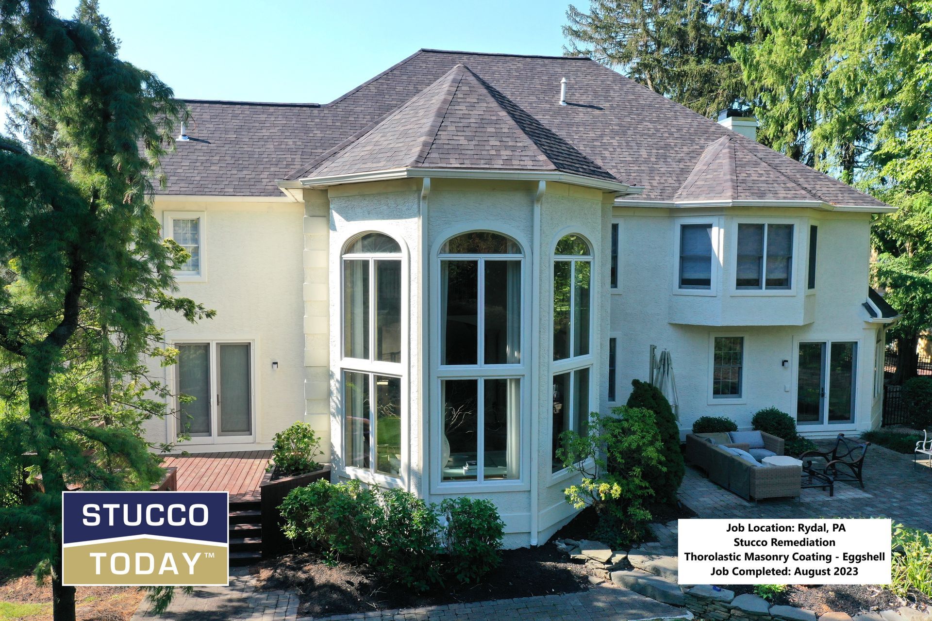 Rear view of a light-colored stucco house with a dark gray roof, lush greenery, and a patio with seating.