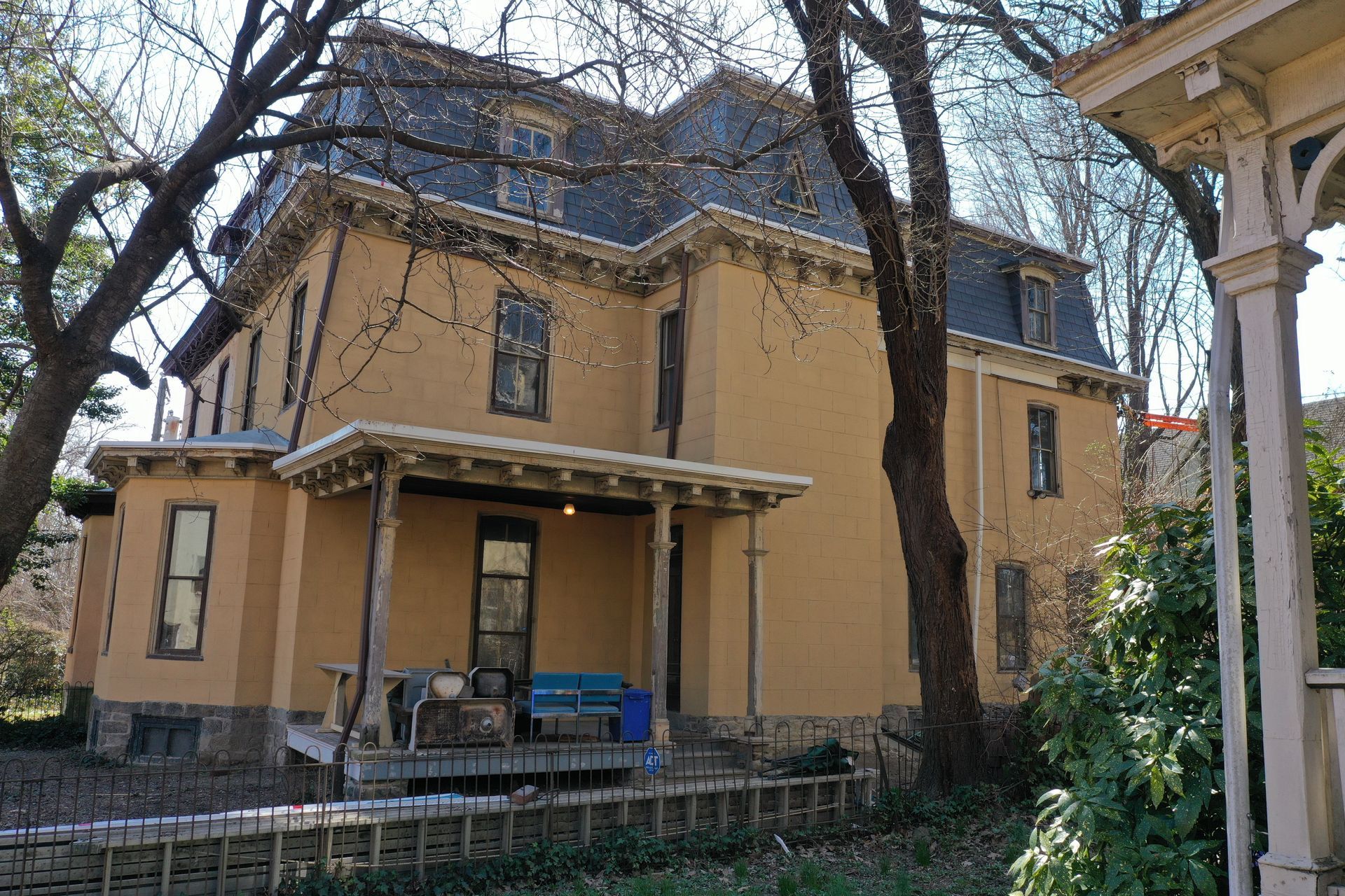 Yellow multi-story house with a mansard roof and porch; trees in the foreground.