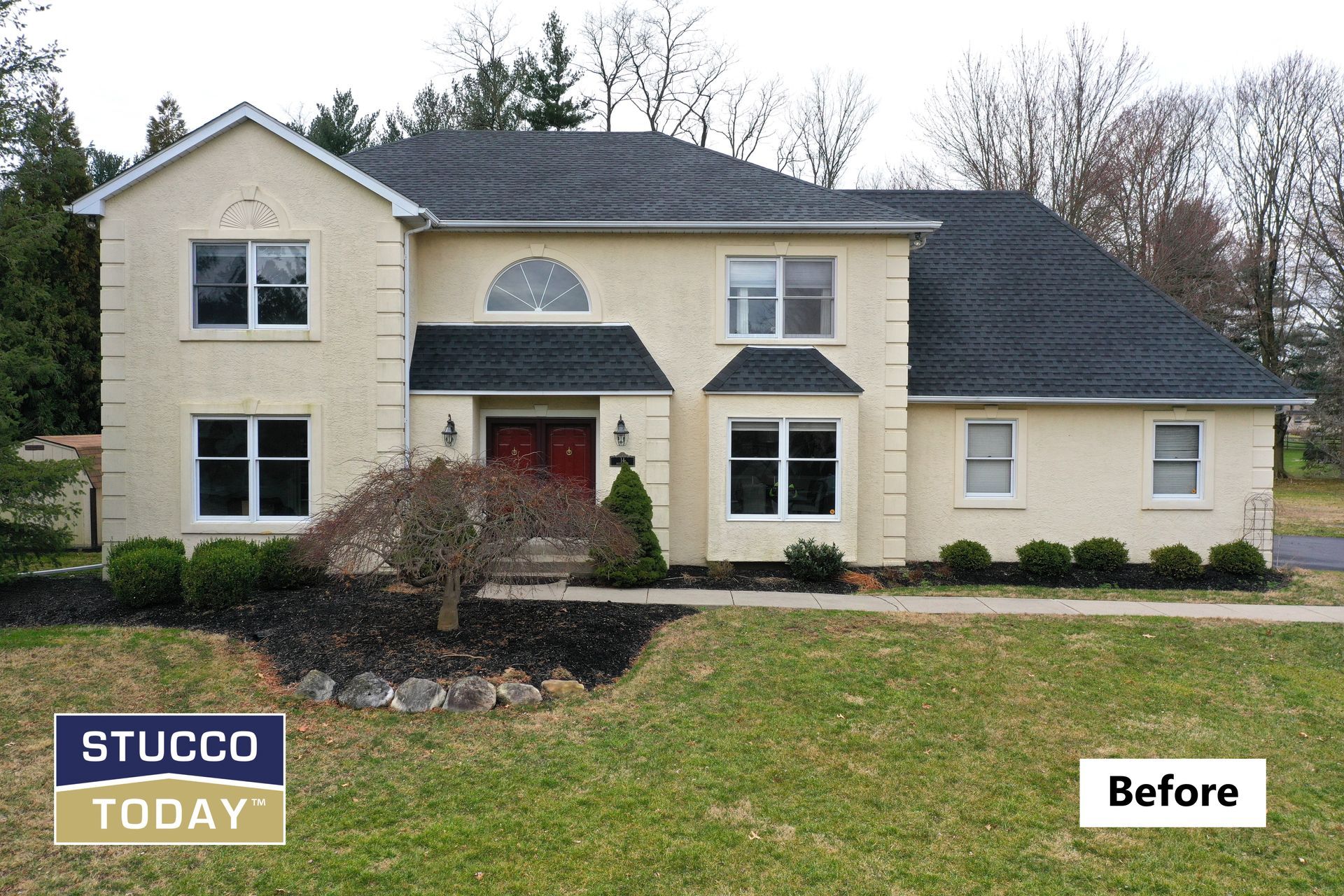 Two-story beige stucco house with dark roof and trim, before stucco repair. Green lawn and shrubs in front.