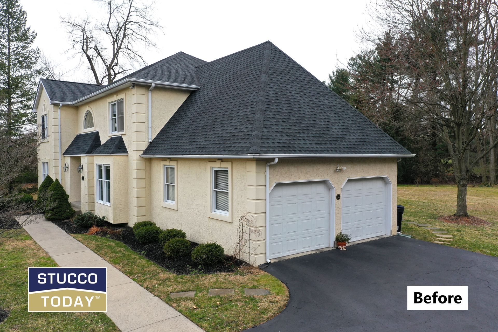 Tan stucco house with black roof and two-car garage, asphalt driveway, before picture.