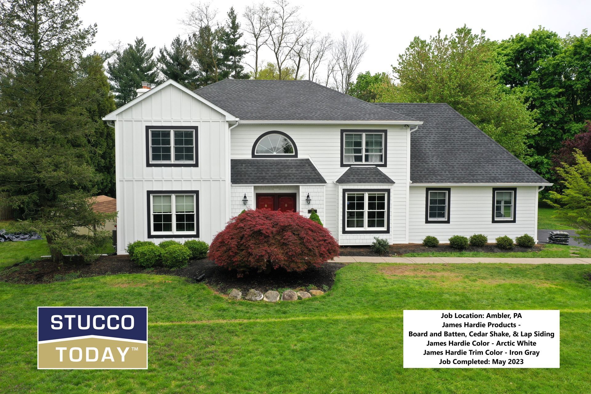 White house with black trim, dark roof. Red door, small bush. Green lawn, trees in background.
