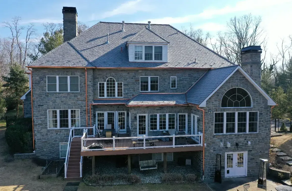 Large stone house with a slate roof, deck, and multiple windows; trees in the background.