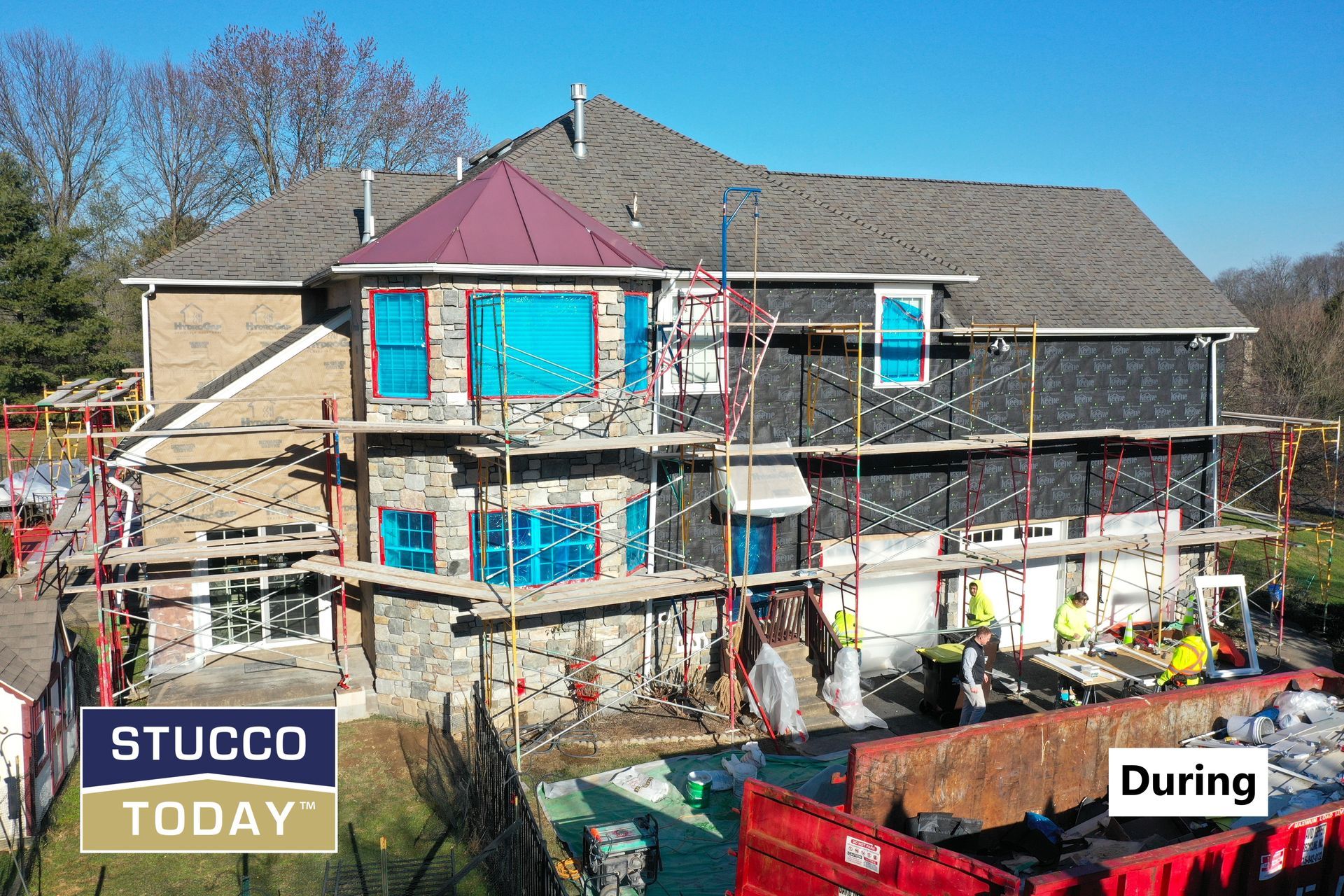 House under construction; scaffolding, stone and dark siding, blue window coverings, workers and dumpster present.