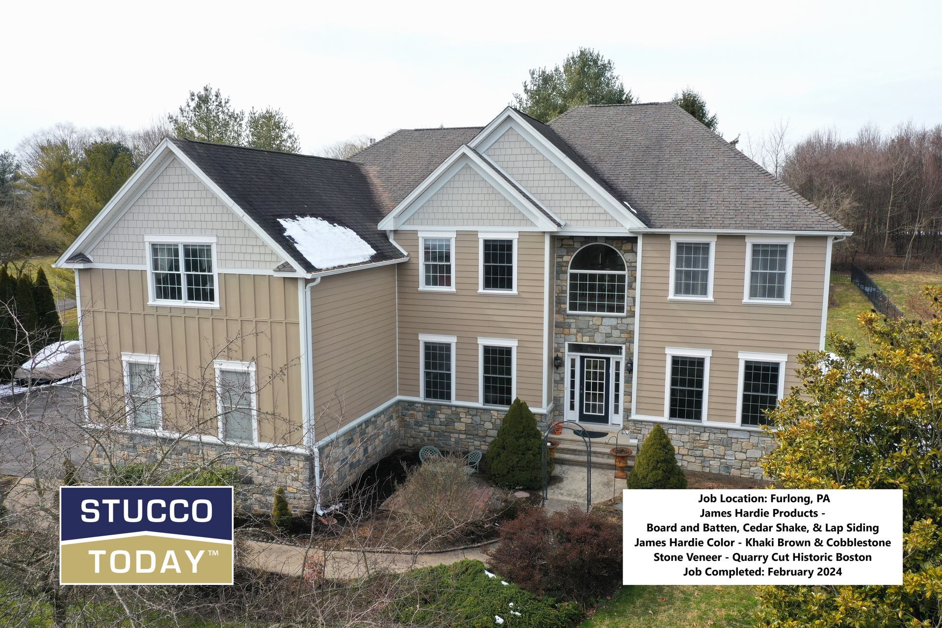 Two-story house with tan siding, stone accents, and snow on the roof.